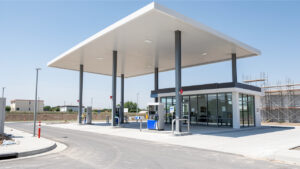 Vue extérieure réaliste d’une station-service moderne en construction, avec accès routier, auvent à structure métallique et zone boutique vitrée.