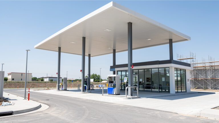 Vue extérieure réaliste d’une station-service moderne en construction, avec accès routier, auvent à structure métallique et zone boutique vitrée.